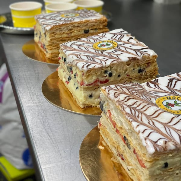 Three Mocha Cafe mille-feuille pastries with white icing, chocolate drizzle, and fruit on gold plates.